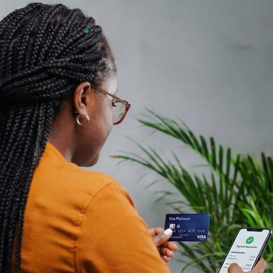 A person in an orange shirt holds a Visa Platinum card and a smartphone displaying a "Payment Successful" message, with a green plant in the background.
