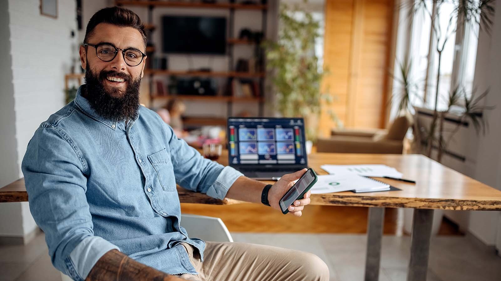Imagem exibe homem sorridente segurando smartphone sentado em escritório.