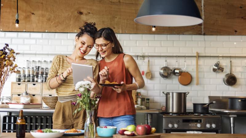 Duas mulheres sorrindo e olhando o seu iPad numa cozinha espaçosa